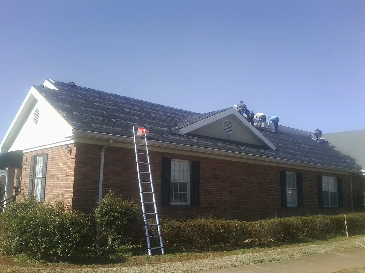 Crew tearing shingles off a brick ranch roof with a ladder set — Emergency Roof Repair work by our crew in Estero